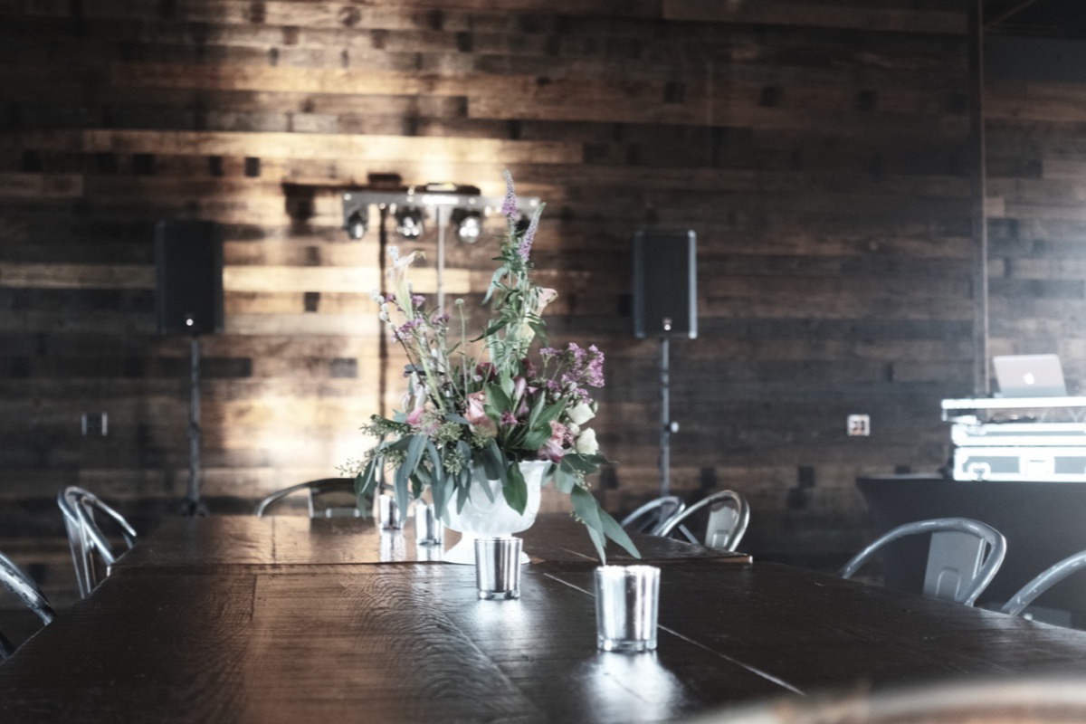 A reception room with a reclaimed wood wall, speakers on stands and a long table set with flowers.