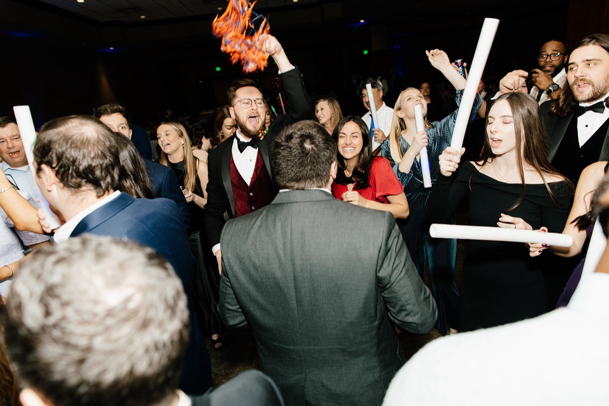 A crowded reception dance floor with guests waving foam glow sticks and confetti in the air.