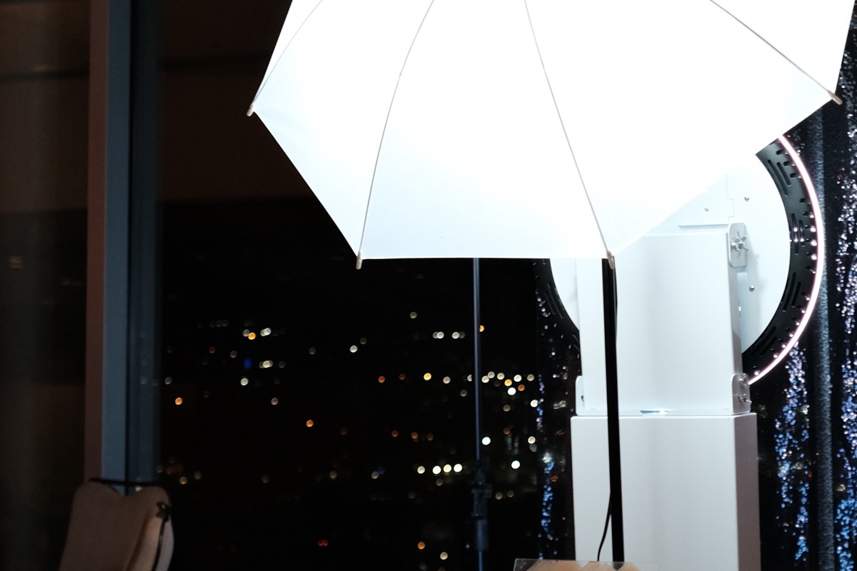 An open-air photo booth lit by an umbrella softbox and a ring light against a sequin backdrop, city lights through the window behind.