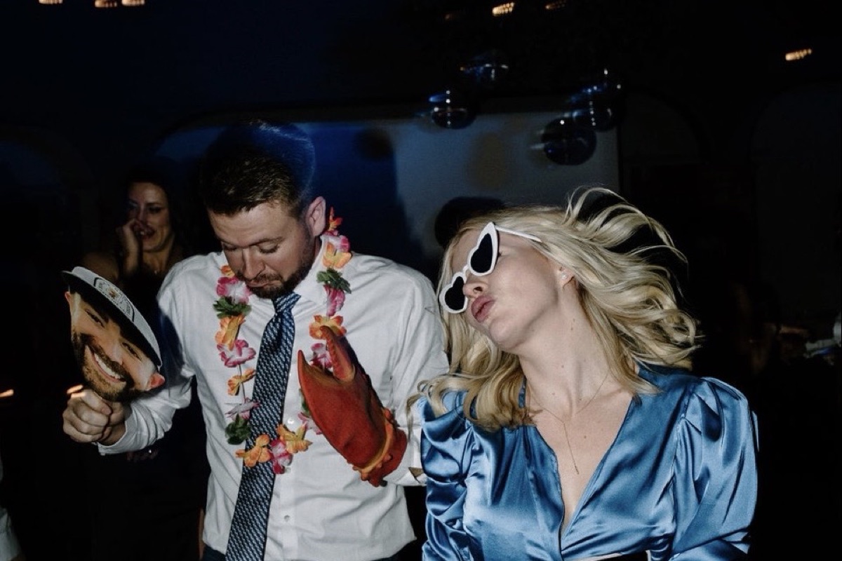 Two guests dancing with photo booth props, one holding a face cutout, the other in heart sunglasses.