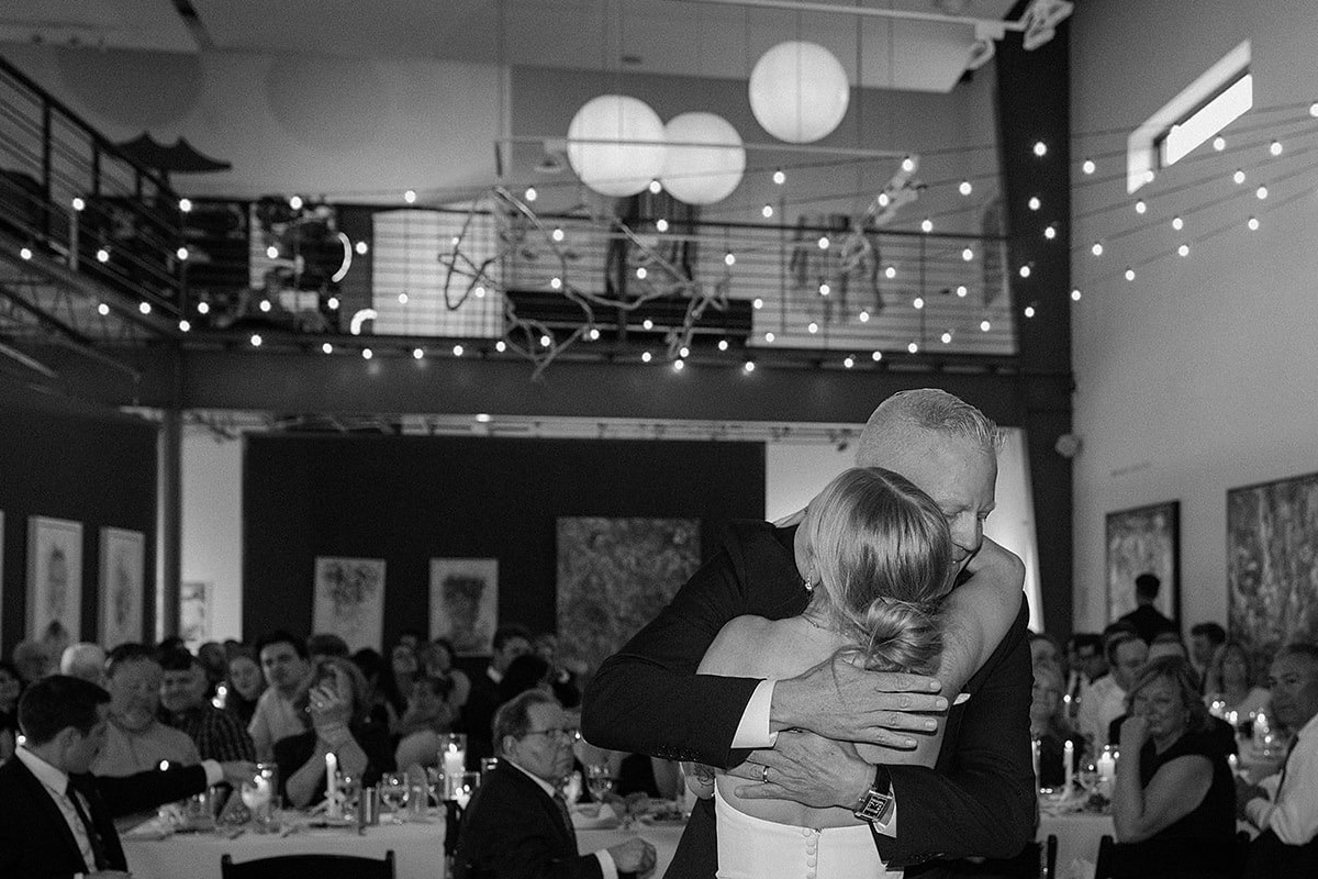 A parent dance in a two storey gallery room, a wall of canvases, a mezzanine and string lights behind, in black and white.