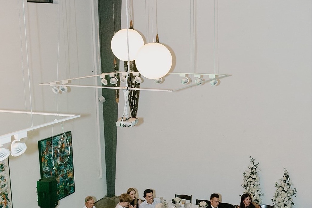 A gallery reception seen from the mezzanine, a sculptural pendant fixture over the floor and guests seated at tables below.