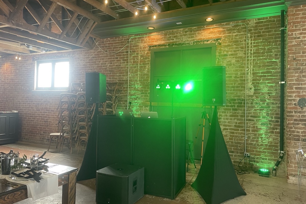 A DJ booth and speakers lit green against an exposed brick wall in an empty room before doors.