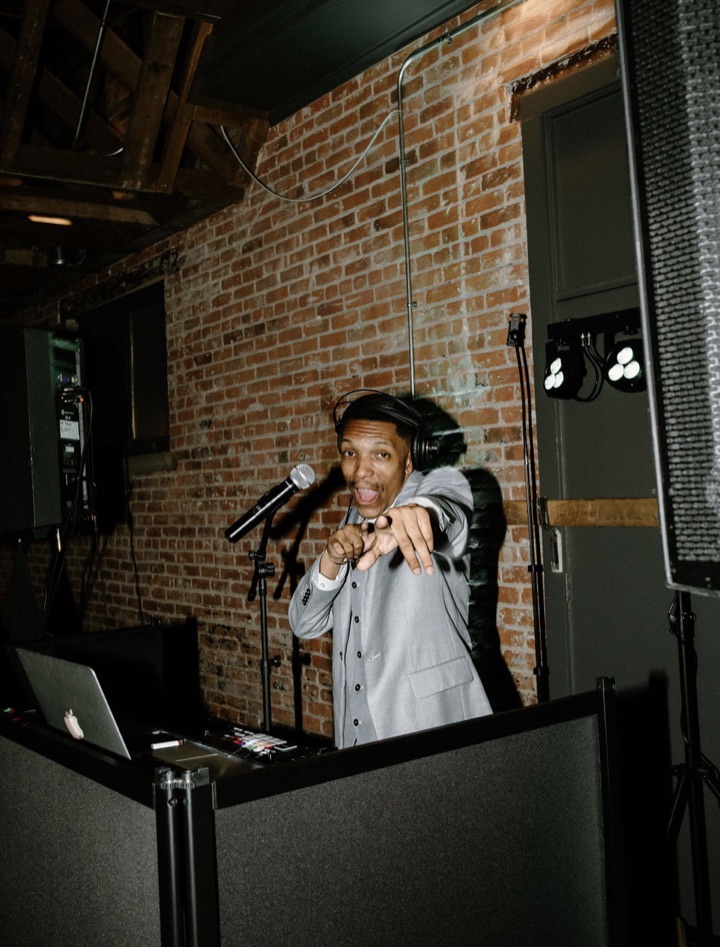 A DJ in a gray suit and headphones pointing at the camera from behind the booth.