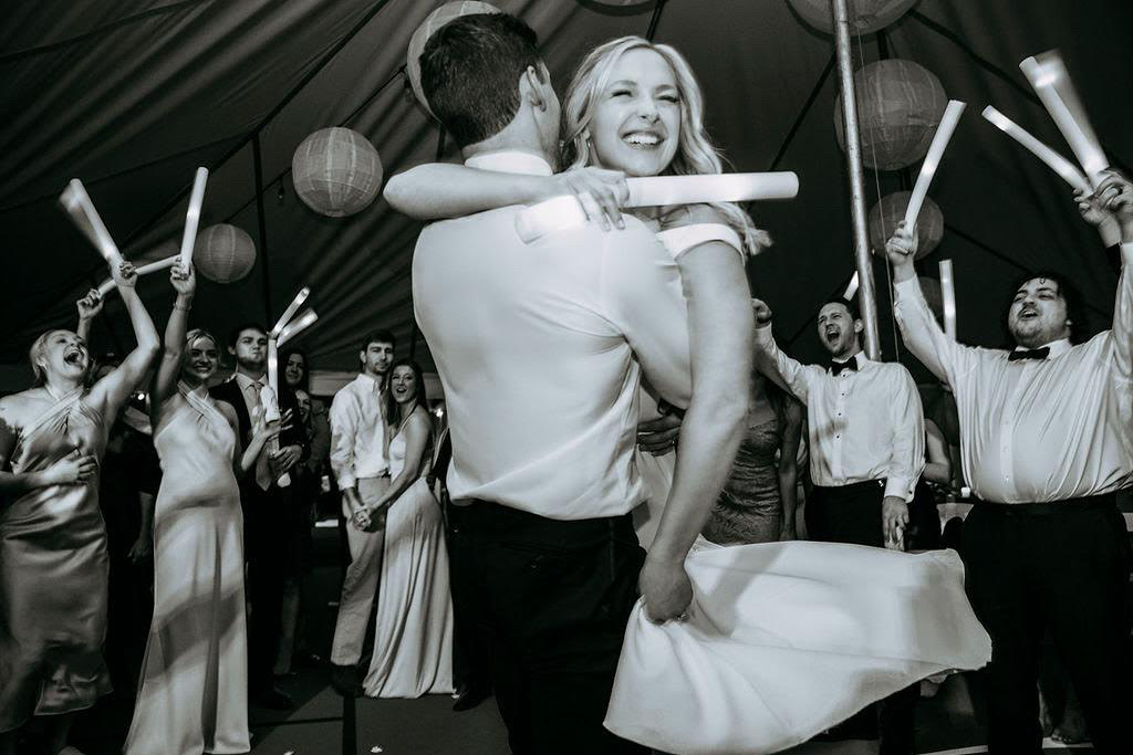 A joyful couple embracing on the dance floor as wedding guests cheer around them, photographed in black and white.