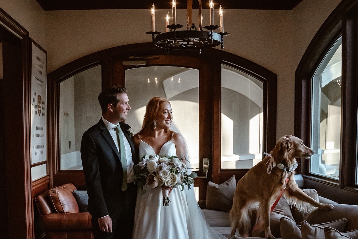 A bride and groom smiling together in an entryway with a golden retriever beside them.
