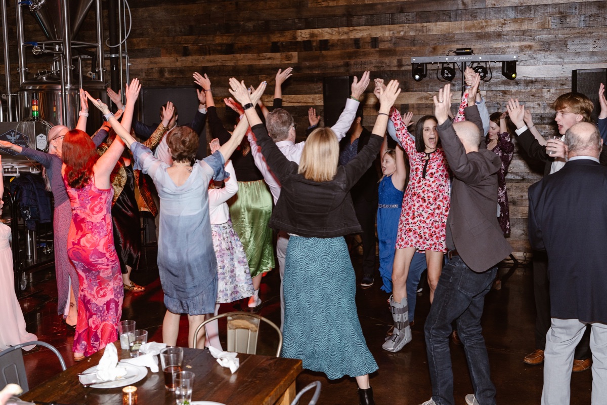 A group of wedding guests raising their arms together on a dance floor at a brewery.